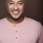 anthonypiresjr_headshot-1-1 A man in front of a brown background wearing a pink shirt and smiling.