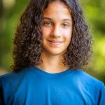 A boy with shoulder-length curly hair wearing a blue t-shirt and smiling.