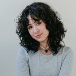 A girl with black hair wearing a black and white necklace and a grey sweater.