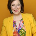 A woman smiling in front of a yellow background wearing a yellow suit.