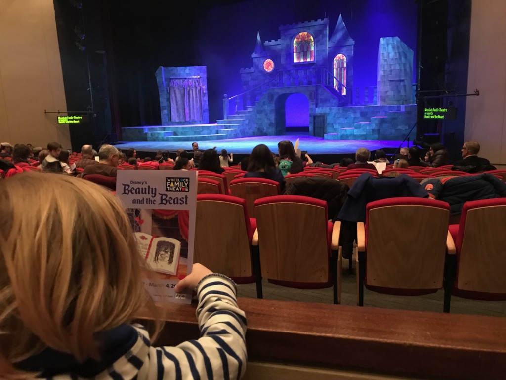 Girl seats in red seats holding Beauty & the Beast program in front of lavish set