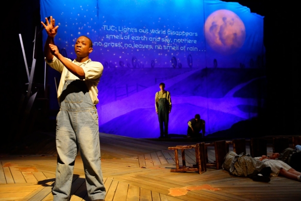 Man stands in overall signing. Woman stands behind. Blue screen is in background with large moon overhead.