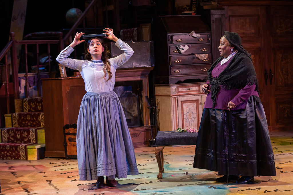 An actor wearing a gray dress holds a book over their head, and another actor in a red and black dress watches from aside.