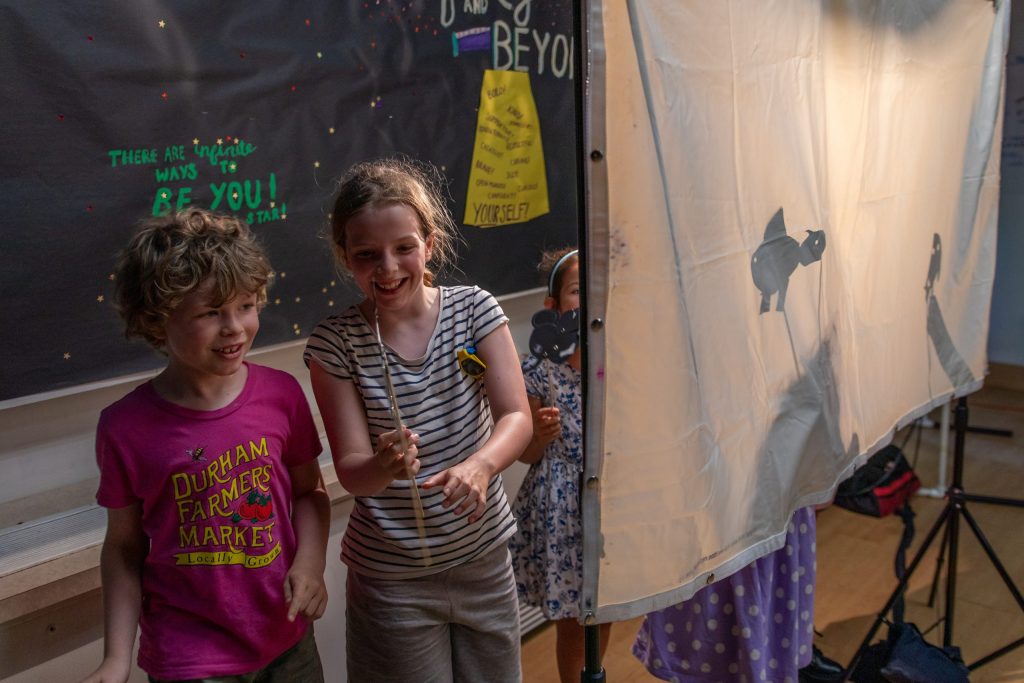 In a theatre demonstration, two young students, one wearing a pink shirt and another in a white shirt with black stripes, happily share their small animal puppets. To the right, two other students use small animal puppets behind a shadow puppet screen.