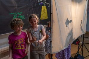 In a theatre demonstration, two young students, one wearing a pink shirt and another in a white shirt with black stripes, happily share their small animal puppets. To the right, two other students use small animal puppets behind a shadow puppet screen.