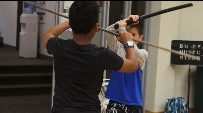 Students with plastic objects fight each other. One student is striking the other's weapon as they defend from the attack.