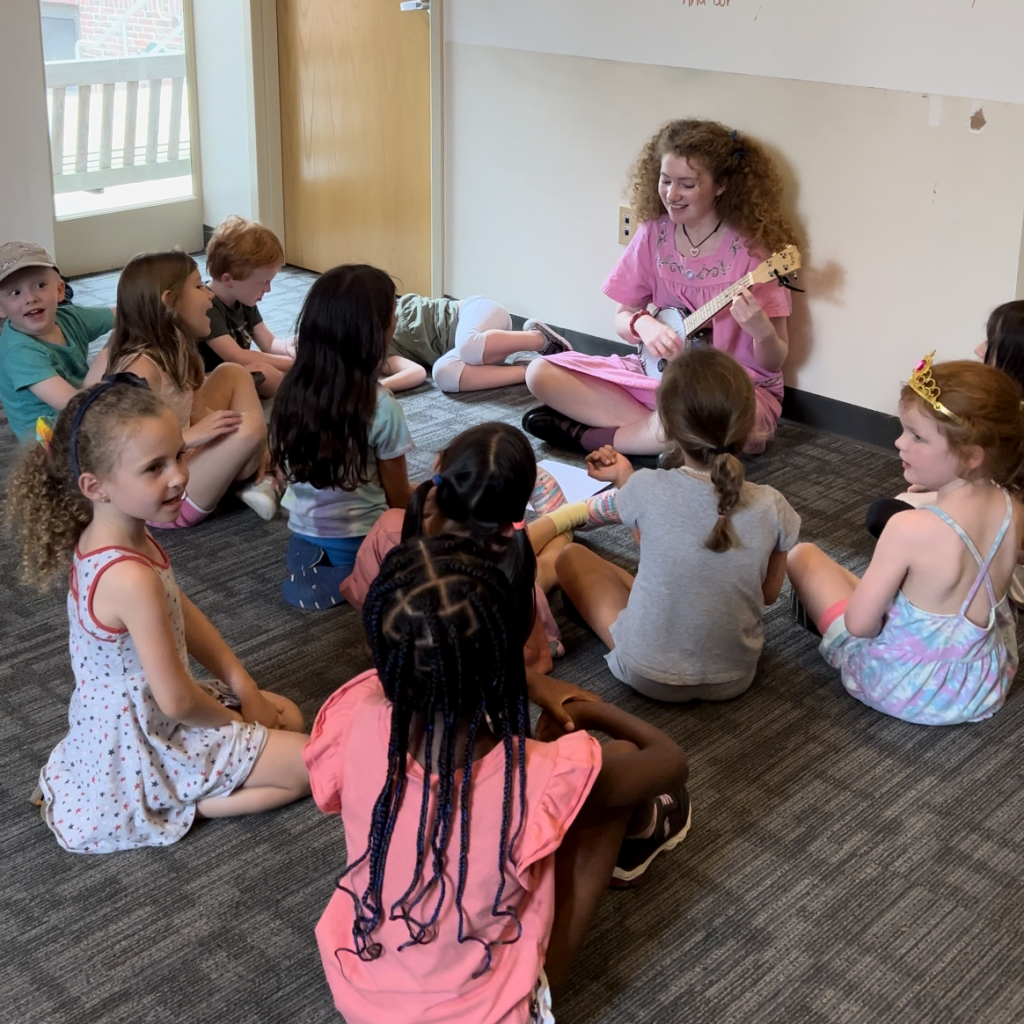 Students sit around a teacher playing the ukulele.