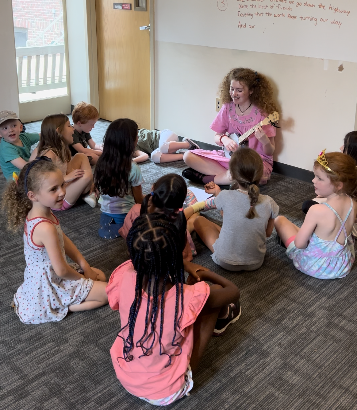 Students sit around a teacher playing the ukulele.