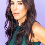 A woman wearing green with brown hair in front of a purple background.