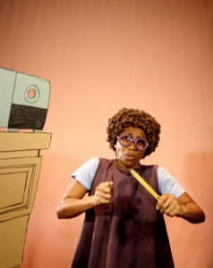 A performer stands against a soft, warm peach-colored backdrop, appearing visibly anxious or startled. They wear round, purple eyeglasses and a wig of tight brown curls, with eyebrows raised and lips slightly parted in a worried expression. Clad in a loose dark-purple pinafore dress over a pale blue t-shirt, they clutch a yellow wooden ruler in both hands—one hand grips it like a baton, the other tenses around it. To the left, a cardboard cut-out fireplace or mantel edge and what appears to be a retro radio or speaker sit partially in frame, reinforcing a stylized, handmade stage set common in Manual Cinema’s puppet-theater aesthetic.