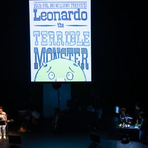 A dimly lit theater stage features a large vertical screen at center stage. The screen displays stylized text reading “Your pal Mo Willems presents Leonardo the Terrible Monster,” along with the top half of the friendly-looking green monster’s face. In the bottom left of the stage, a performer sits on a chair, playing an acoustic guitar while illuminated by soft spotlight. To the right, two other performers work at a table under focused lighting, apparently manipulating props or overseeing visuals. The surrounding stage is mostly dark, emphasizing the performers and the screen’s warm glow.