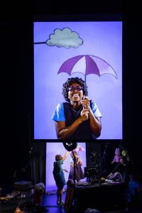 A theatrical scene unfolds on a dimly lit stage featuring a large vertical screen dominating the background. On the screen, a close-up of a smiling performer holding a pink-and-white umbrella is visible—she wears round purple glasses, short curly hair, and a navy dress over a light blue shirt. Above her on the screen is a whimsical gray cloud prop affixed to a rod. In front of the screen, the same performer appears in person, holding the umbrella, while another performer stands beside her, adjusting the cloud overhead. To the right, a third stagehand or performer crouches at a table filled with puppets, props, and lighting equipment. The overall scene showcases the creative mechanics of a puppet-theater performance, where projected imagery, live manipulation, and playful props combine to create a magical theatrical moment.