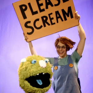A cheerful stage performer stands center frame against a glowing lavender backdrop, holding a large, hand-made cardboard sign overhead that reads “PLEASE SCREAM” in bold uppercase letters. The performer wears round glasses, a short, spiky orange wig, and a blue jumper dress over a light green shirt, their expression bright and inviting. Below them is a fuzzy, lime-green monster puppet with wide blue eyes, a round pink nose, and an open mouth showing a blue tongue—its expression joyful and a bit goofy. The ensemble creates a playful, interactive moment that invites audience engagement.