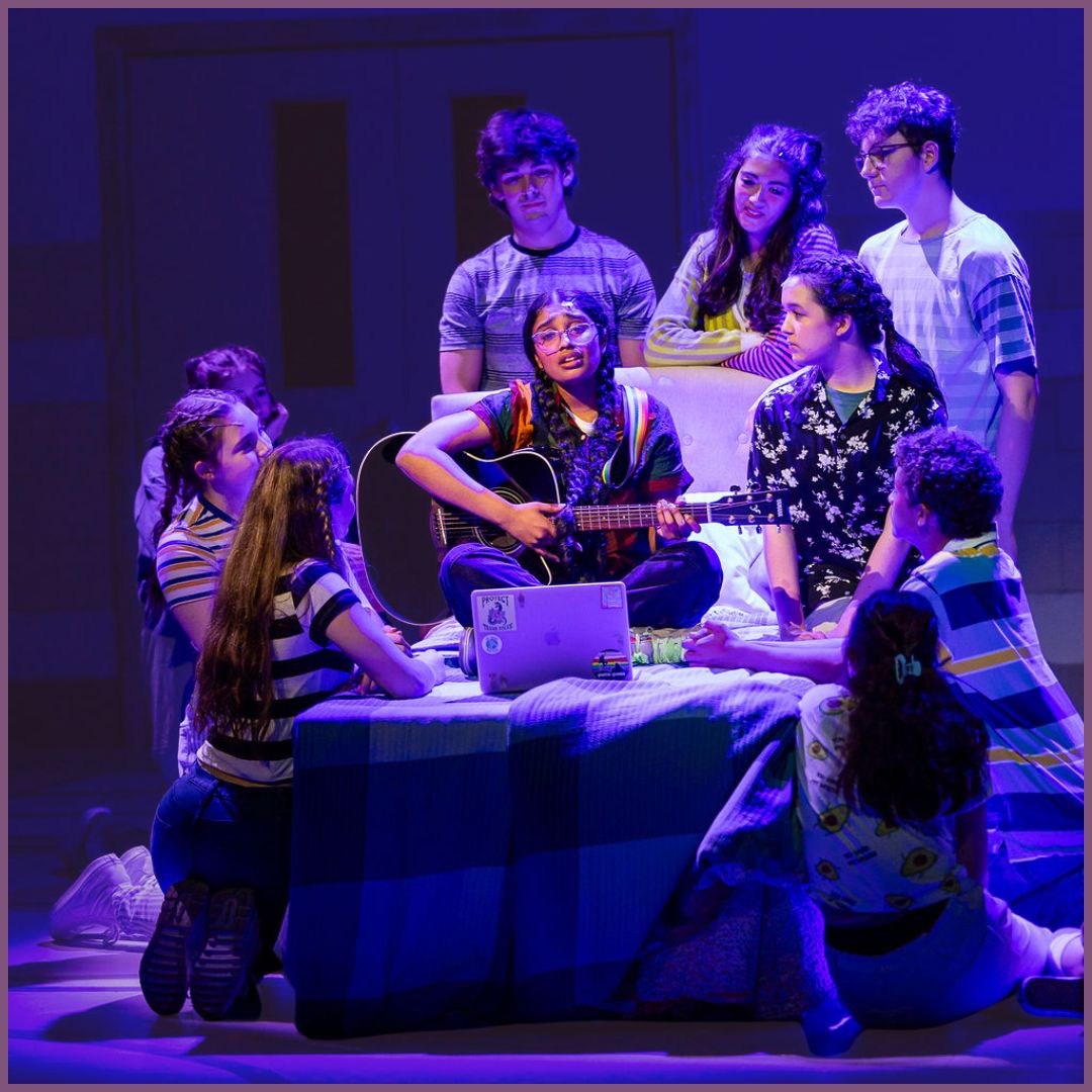 A girl with long braids sits on a bed holding a guitar surrounded by a group of teenagers.