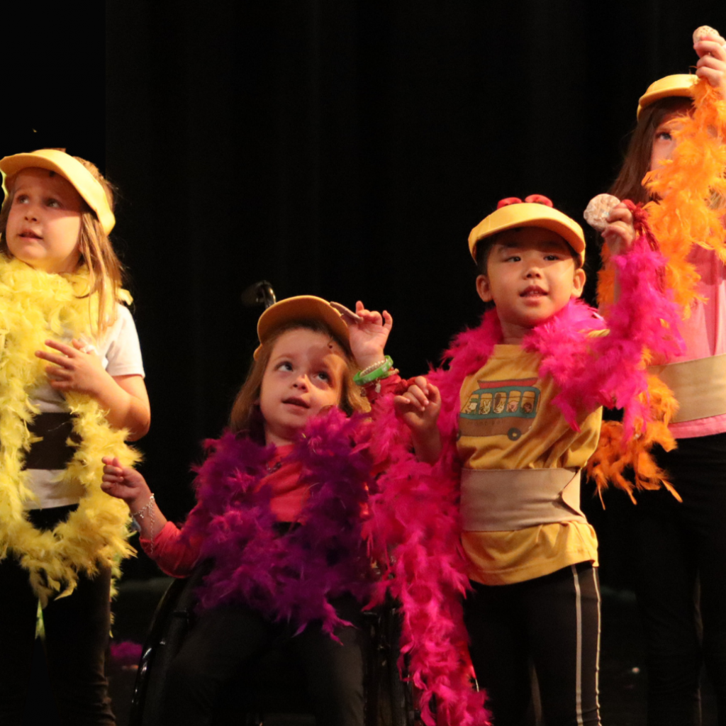 frog-and-toad-2026-education-main-page Four students wearing yellow vizors and multicolored boas point to the sky.