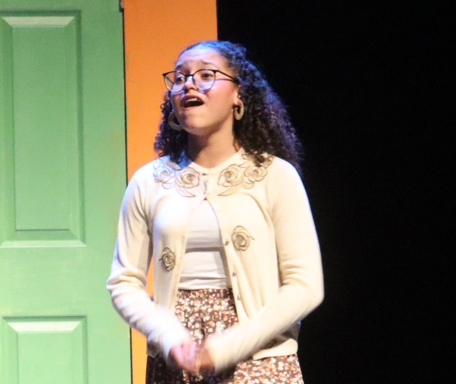 matilda-2025-education-landing-page A student in a white cardigan and brown knee length skirt sings a sad song.