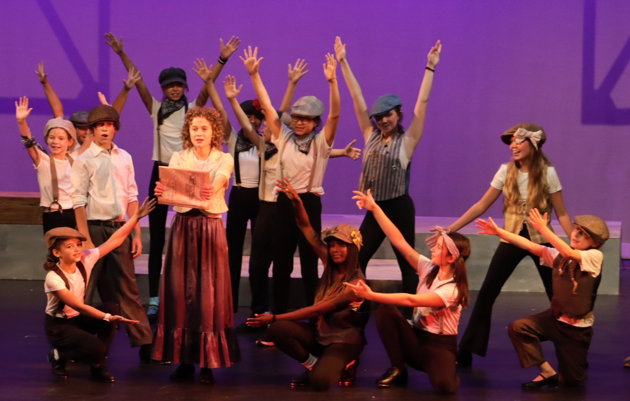 newsies-2025-image-4673 Students frame a performer holding a newspaper in a energetic pose