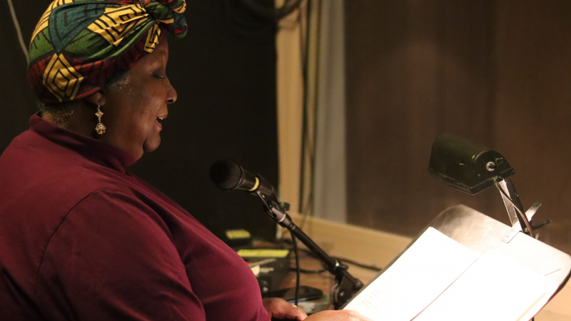 A Black woman in a colorful headscarf reads from a script into a microphone.