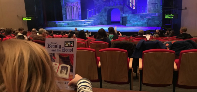 Girl seats in red seats holding Beauty & the Beast program in front of lavish set